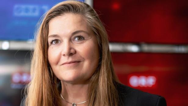 Bettina Parschalk mit langem, braunem Haar trägt ein schwarzes Sakko vor Bildschirmen mit dem ORF-Logo.