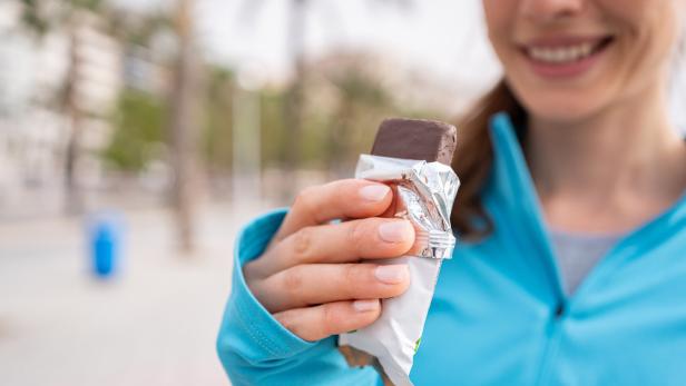 Lächelnde Frau in blauer Jacke hält einen ausgepackten Schokoriegel in der Hand.