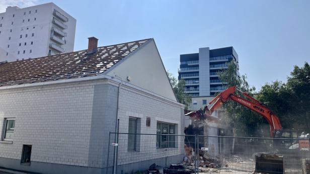 Bagger reißt ein Gebäude neben einem weißen Haus ab.