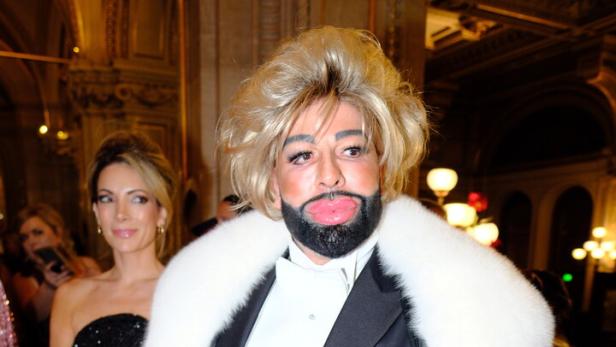 Harald Glööckler trägt eine blonde Perücke, einen schwarzen Anzug mit weißem Pelzkragen und hält eine kleine Tasche in den Händen.
