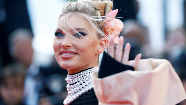Elsa Hosk lächelt und winkt in die Kamera, geschmückt mit einer auffälligen Perlenkette und einer Blume im Haar.