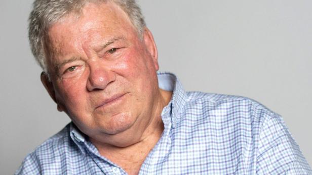 William Shatner trägt ein hellblau kariertes Hemd und blickt vor einem neutralen Hintergrund in die Kamera.