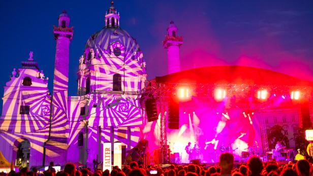 Die Wiener Karlskirche wird bei einem Open-Air-Konzert mit Lichtprojektionen in Pink und Violett angestrahlt, während eine Band auf der Bühne spielt.