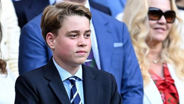 Prinz George trägt einen dunklen Anzug mit Krawatte und stützt sich auf eine hölzerne Brüstung.