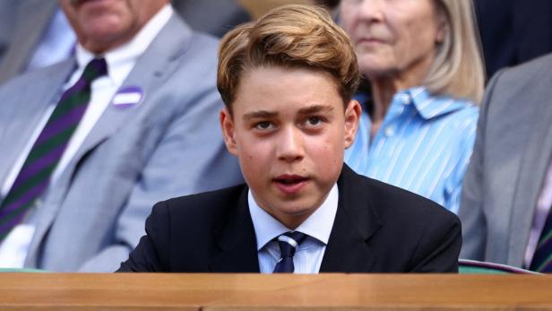 Prinz George sitzt in einem dunklen Anzug mit Krawatte auf der Tribüne, während im Hintergrund weitere Personen zu sehen sind.
