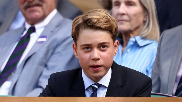 Prinz George sitzt in einem dunklen Anzug mit Krawatte auf der Tribüne, während im Hintergrund weitere Personen zu sehen sind.