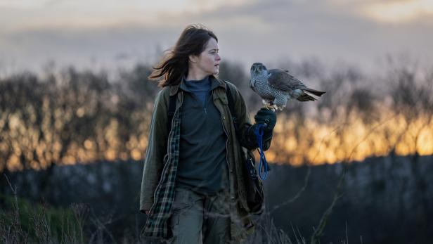 Eine Frau in Outdoor-Kleidung (Claire Foy) trägt einen Greifvogel auf der behandschuhten Hand in einer Landschaft bei Sonnenuntergang.