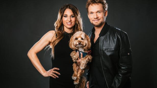 Eine Frau in einem schwarzen Kleid und ein Mann in einer Lederjacke halten gemeinsam einen kleinen, hellbraunen Hund vor einem dunklen Hintergrund.
