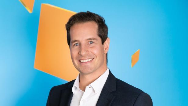 Geschäftsführer Philipp König lächelt, im Anzug vor blauem Hintergrund mit orangefarbenen Flächen, über gute Zahlen der kronehit-Senderfamilie.