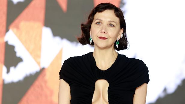 Maggie Gyllenhaal trägt ein schwarzes Kleid mit Cut-out und auffällige grüne Ohrringe vor einem unscharfen Hintergrund.