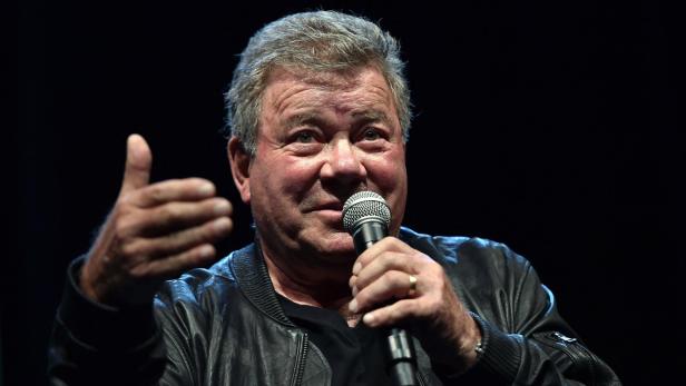 William Shatner spricht vor einem dunklen Hintergrund in ein Mikrofon und gestikuliert mit der rechten Hand.