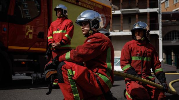 Drei Feuerwehrleute der Sapeurs-Pompiers de Paris in roter Schutzkleidung und mit Helmen knien vor einem Einsatzfahrzeug auf der Straße.