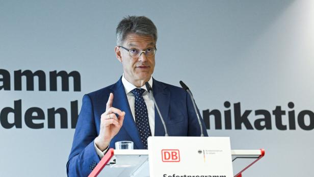 Mann im blauen Anzug spricht am Podium mit erhobenem Finger vor DB-Transparent.