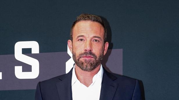 Ben Affleck trägt einen dunkelblauen Anzug mit weißem Hemd vor einer dunklen Hintergrundwand mit Schriftzügen.