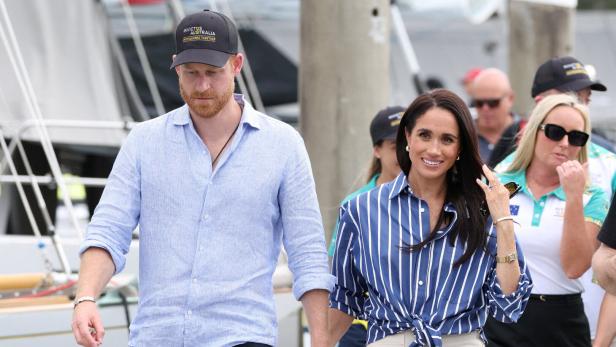 Prinz Harry und Herzogin Meghan gehen händehaltend an einem Hafen entlang, wobei Harry eine Invictus-Australia-Kappe trägt.
