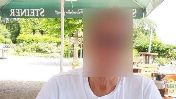 Eine Person sitzt im Freien an einem Tisch unter einem großen Sonnenschirm; das Gesicht ist unkenntlich gemacht.