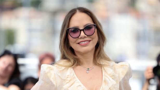 Ornella Muti lächelt in einem hellen, gerüschten Kleid und mit einer getönten Sonnenbrille vor einer unscharfen Kulisse.