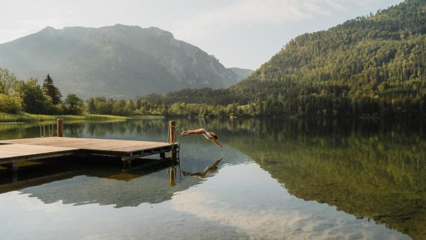 Eine Frau springt von einem Steg in einen stillen Bergsee, umgeben von bewaldeten Hängen und Spiegelungen im Wasser.