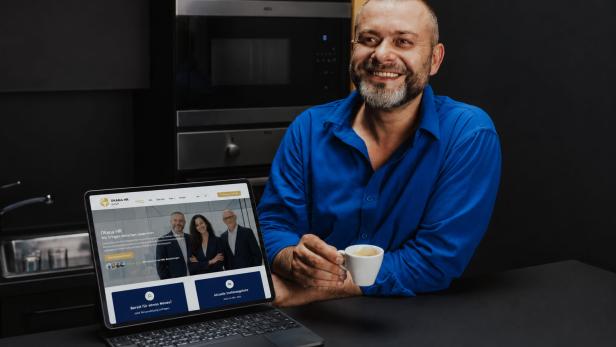 Mann mit Bart und blauem Hemd sitzt an schwarzem Tisch und hält weiße Tasse neben offenem Laptop mit Webseite.