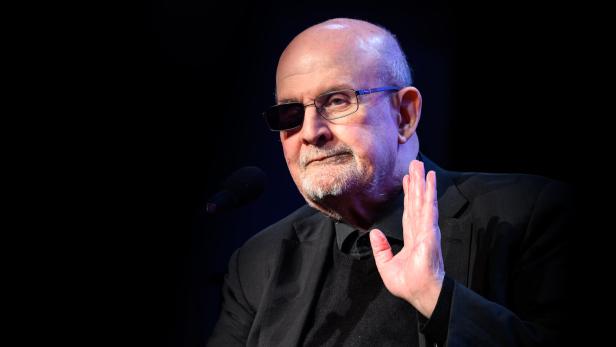 Salman Rushdie mit Brille und Augenklappe, der die Hand hebt, auf einer Bühne.