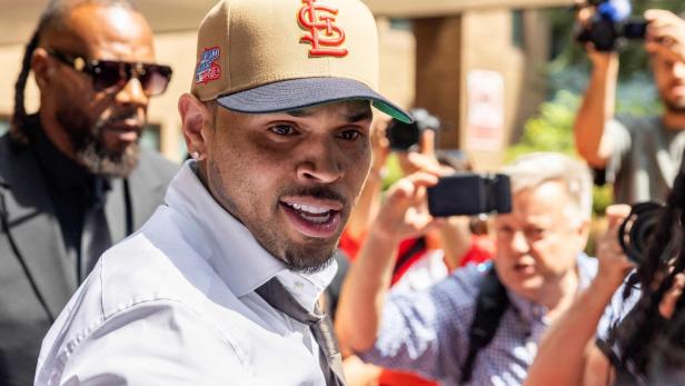 Chris Brown trägt ein weißes Hemd und eine beigefarbene St. Louis Cardinals-Kappe, während er von Fotografen umringt wird.