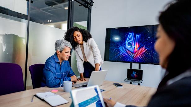 Drei Personen besprechen am Konferenztisch KI, während auf einem Monitor ein leuchtendes „AI“ erscheint.