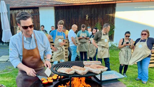 Mann grillt Fleisch und Gemüse an einer offenen Feuerstelle, während mehrere Personen in Schürzen zuschauen.
