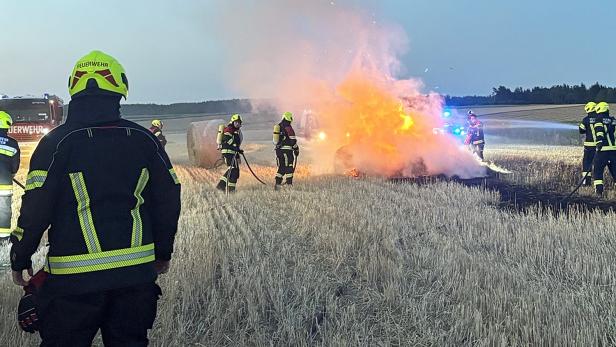 Feuerwehrleute löschen einen brennenden Wagen auf einem Feld, während Rauch und Flammen aufsteigen.