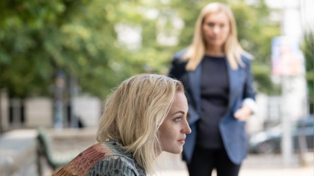 Blonde Frau im Profil im Vordergrund, dahinter steht unscharf eine weitere Frau im Freien.