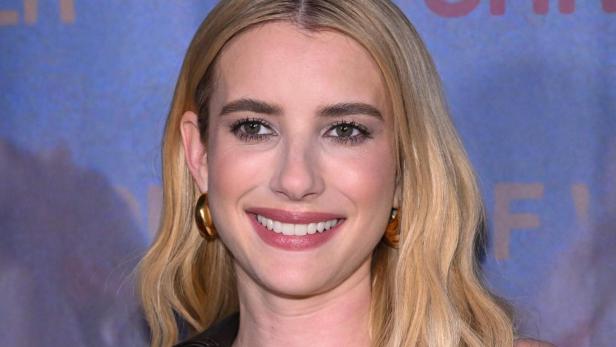 Emma Roberts mit blondem Haar lacht und klatscht inmitten einer jubelnden Menschenmenge bei einer Sportveranstaltung.
