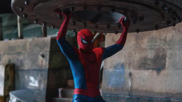 Spider-Man hebt in einem rot-blauen Kostüm eine schwere, metallische Konstruktion über seinen Kopf.