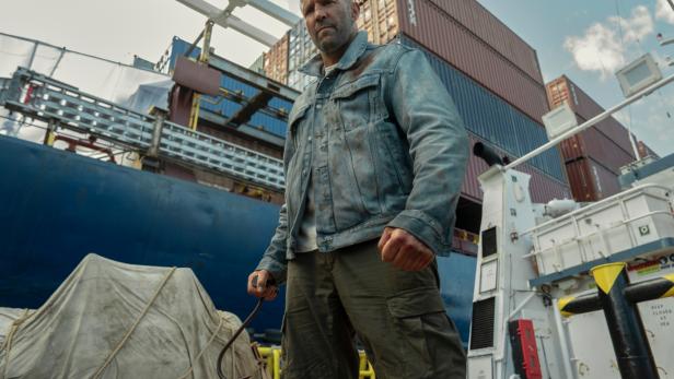 Jason Statham steht mit einem Haken in der Hand vor einem Frachtschiff mit Containern.