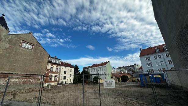 Umzäunte Baustelle zwischen Wohnhäusern unter dramatischem Wolkenhimmel.