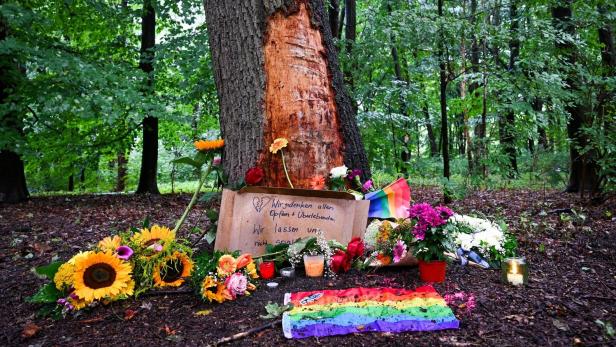 Ein Baum im Wald mit Blumen, Kerzen und einer Regenbogenflagge sowie einem Schild mit der Aufschrift „Wir gedenken allen Opfern & Überlebenden“.