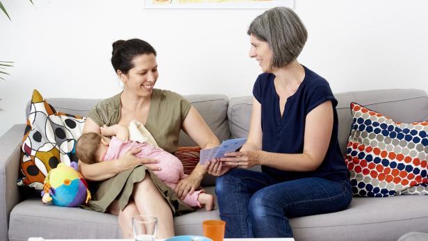 Eine Frau mit einem Säugling sitzt auf einem Sofa, daneben sitzt eine Familienbegleiterin, die ihr Informationsmaterial zeigt.