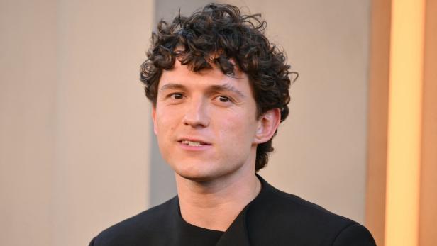 Tom Holland mit lockigem Haar und schwarzem Oberteil vor einem neutralen Hintergrund.
