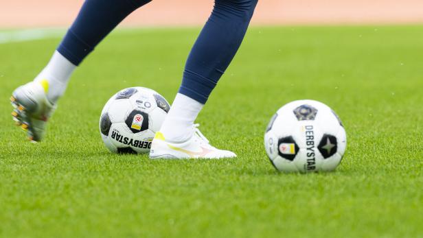 Fußballspiel mit zwei Bällen und den Beinen einer Spielerin auf dem Rasen.