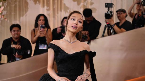 Ariana Grande trägt ein schwarzes schulterfreies Kleid und eine Halskette, während sie von Fotografen umgeben ist.