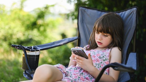 Ein Mädchen sitzt in einem Campinghocker und tippst auf einem Smartphone.