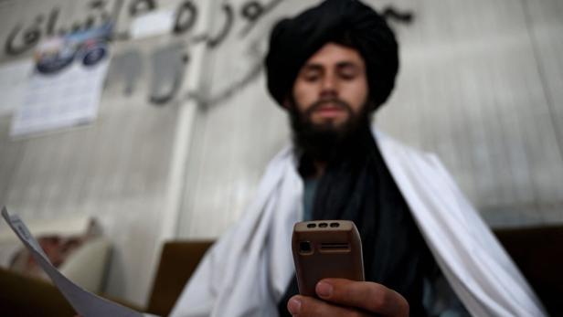 Ein bärtiger Mann mit Turban hält ein Handy und ein Papier vor einer Wand mit Schriftzug.