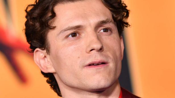 Tom Holland trägt ein dunkelrotes Sakko mit einem roten Hemd und einer dunklen Krawatte vor einem orangefarbenen Hintergrund.