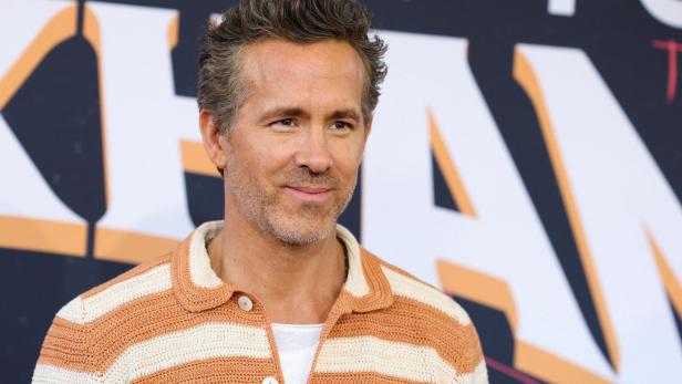 Ryan Reynolds lächelt vor einem Hintergrund mit großen Buchstaben in die Kamera.