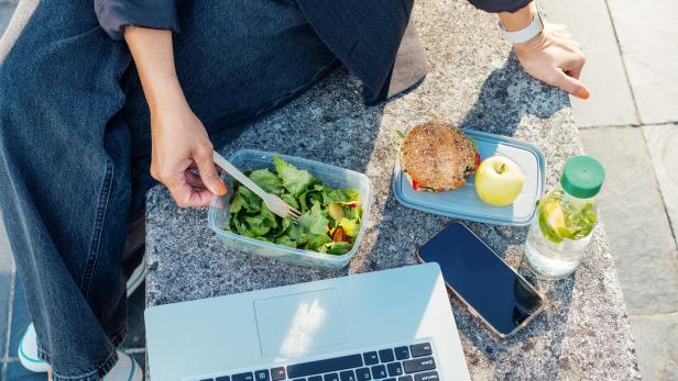 Top view business woman having lunch break, eating fast and healthy in front of laptop outdoors. Balanced diet lunchbox with fresh salad, sandwich, apple, water. Healthy eating habits and well-being.