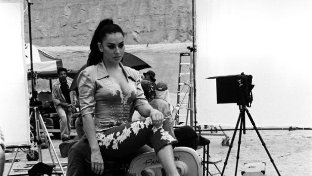 Eine Frau sitzt auf einer Filmkamera am Set, umgeben von Lichtreflektoren und Crew.