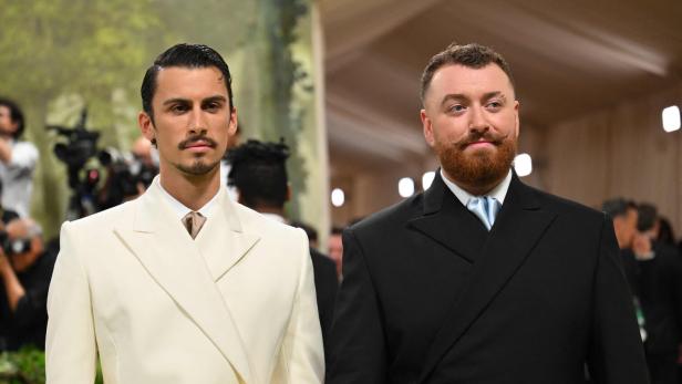 Sam Smith und Christian Cowan in eleganten Anzügen mit goldenen Rosen-Anstecknadeln bei einer Veranstaltung.