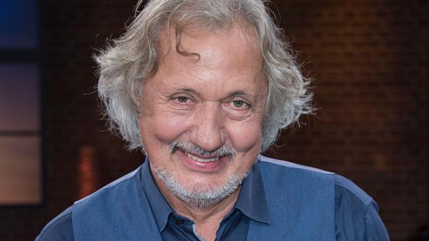 Konrad Beikircher lächelt in blauer Weste vor einer Studiokulisse.