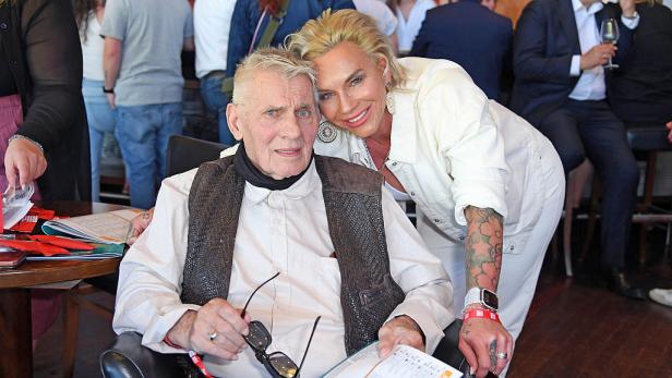 Heinz Hoenig im Rollstuhl mit seiner Frau Annika Kärsten-Hoenig  im weißen Outfit im Hintergrund.