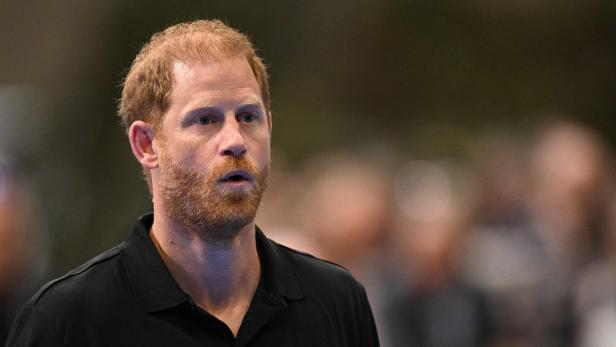 Prinz Harry trägt ein schwarzes Poloshirt mit dem Logo der Invictus Games Foundation und blickt mit leicht geöffnetem Mund zur Seite.