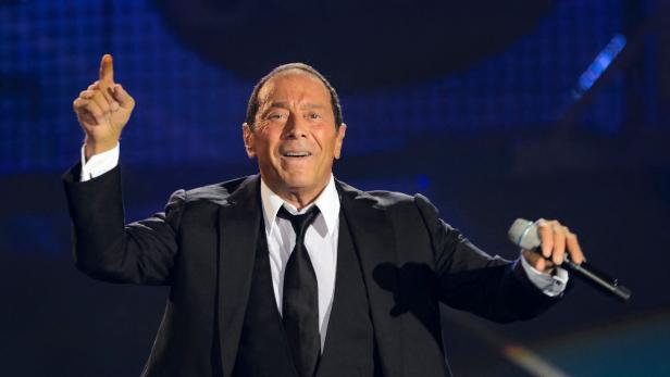 Paul Anka singt in einem schwarzen Anzug mit Krawatte auf einer Bühne und hebt dabei einen Finger in die Luft.