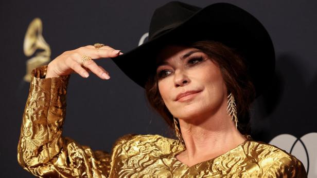 Shania Twain trägt einen schwarzen Cowboyhut und ein goldenes, gemustertes Oberteil vor einem dunklen Hintergrund.
